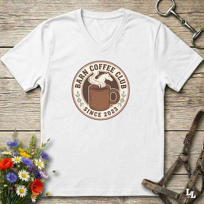 A white v-neck t-shirt with a circular 'Barm Coffee Club Since 2023' graphic on the chest, displayed in a flat-lay arrangement on a rustic wooden background with equestrian accessories and flowers.