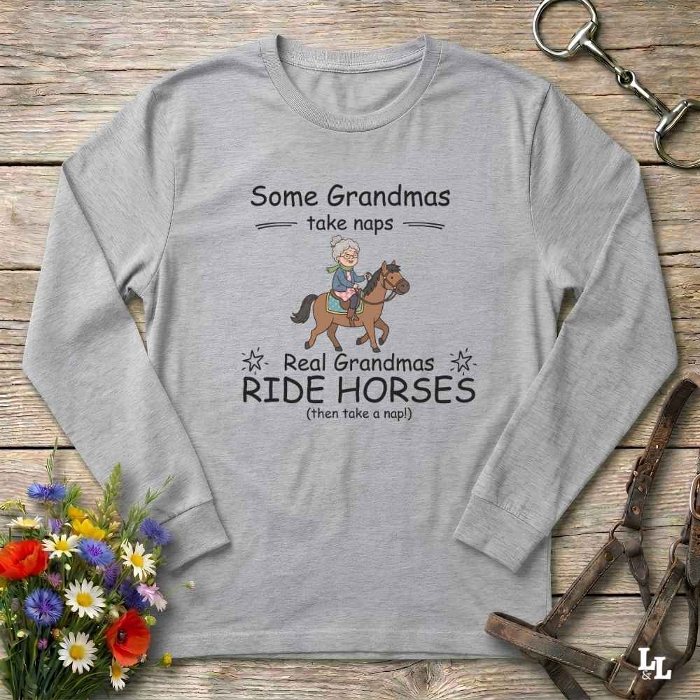 A light gray long-sleeve t-shirt with a graphic print of a grandma riding a horse, displayed on a rustic wooden background with equestrian accessories and flowers.