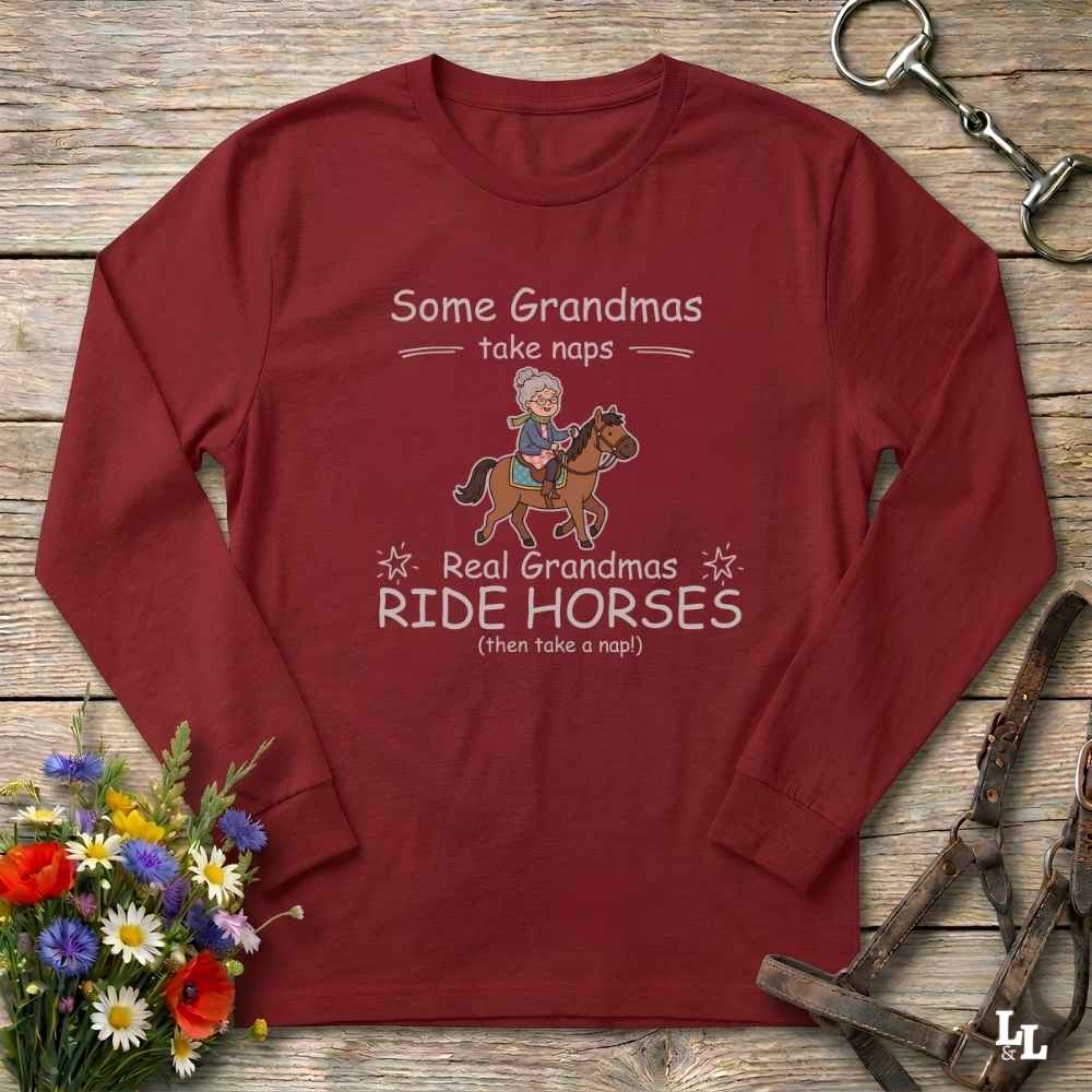 A long-sleeve crewneck t-shirt in maroon red, laid flat on a rustic wooden background. The shirt features a graphic print with the text 'Some Grandmas take naps' and 'Real Grandmas RIDE HORSES (then take a nap!)' surrounding an illustration of a grandma riding a horse.