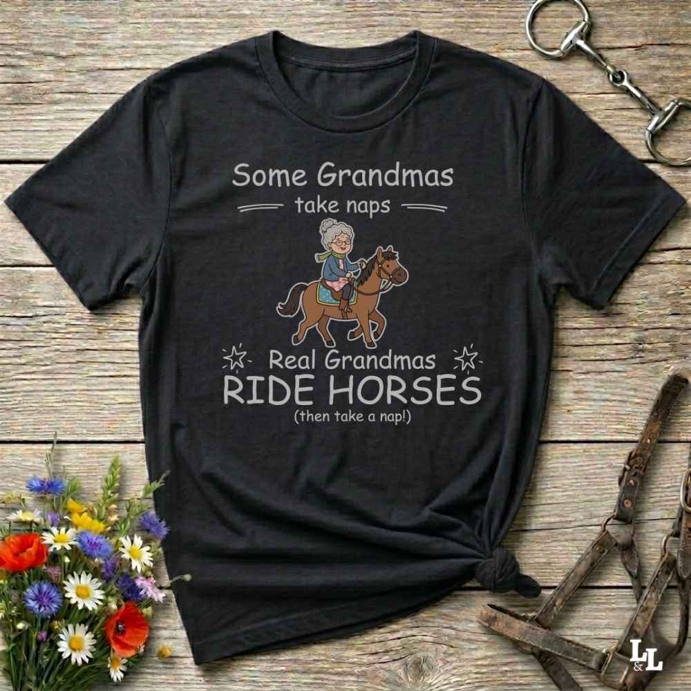 A black graphic t-shirt laid flat on a rustic wooden background, styled with equestrian accessories and flowers. The shirt features a cartoon illustration of a grandma riding a horse and the text 'Some Grandmas take naps Real Grandmas RIDE HORSES (then take a nap!)'.