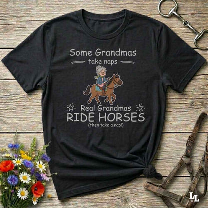 A black graphic t-shirt laid flat on a rustic wooden background, styled with equestrian accessories and flowers. The shirt features a cartoon illustration of a grandma riding a horse and the text 'Some Grandmas take naps Real Grandmas RIDE HORSES (then take a nap!)'.