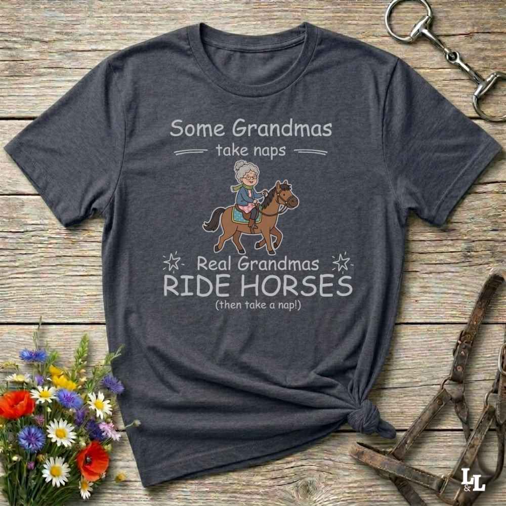 A dark heather gray graphic t-shirt laid flat on a rustic wooden surface, styled with a small bouquet of flowers and horse tack props. The shirt features a cartoon illustration of a grandma riding a horse and the text 'Some Grandmas take naps' and 'Real Grandmas RIDE HORSES (then take a nap!)'.
