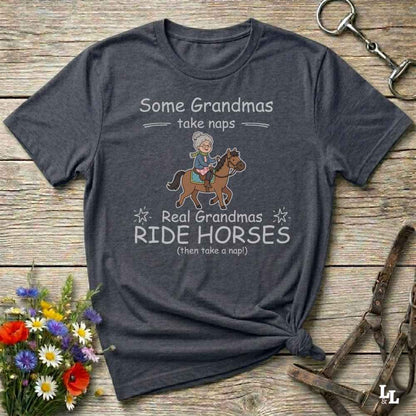 A dark heather gray graphic t-shirt laid flat on a rustic wooden surface, styled with a small bouquet of flowers and horse tack props. The shirt features a cartoon illustration of a grandma riding a horse and the text 'Some Grandmas take naps' and 'Real Grandmas RIDE HORSES (then take a nap!)'.