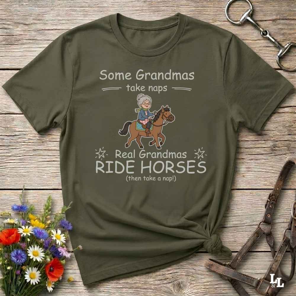 An olive green graphic t-shirt laid flat on a rustic wooden background, styled with equestrian accessories and a small bouquet of flowers. The shirt features a cartoon illustration of a grandma riding a horse and the text 'Some Grandmas take naps Real Grandmas RIDE HORSES (then take a nap!)'.