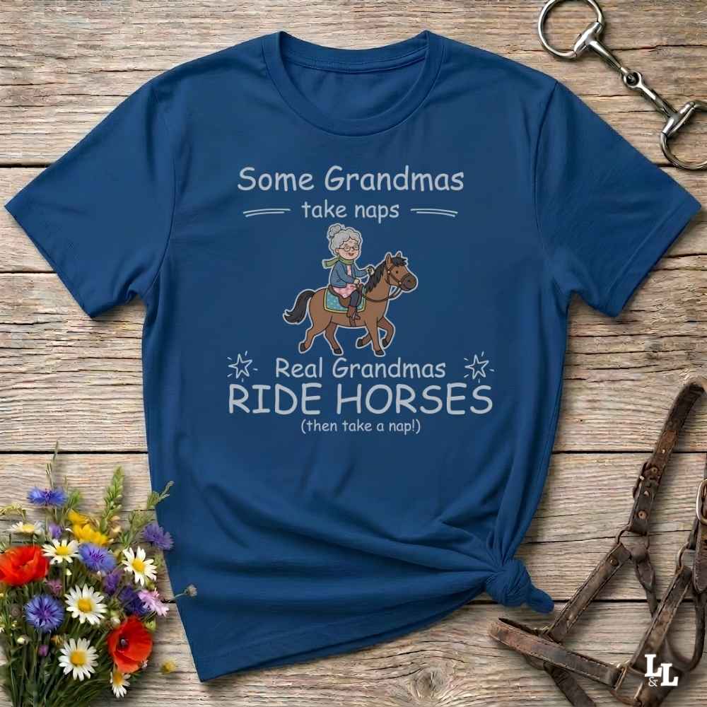 A navy blue graphic t-shirt laid flat on a rustic wooden surface, styled with equestrian accessories and flowers. The shirt features a cartoon illustration of a grandma riding a horse, with the text 'Some Grandmas take naps' and 'Real Grandmas RIDE HORSES (then take a nap!)'.