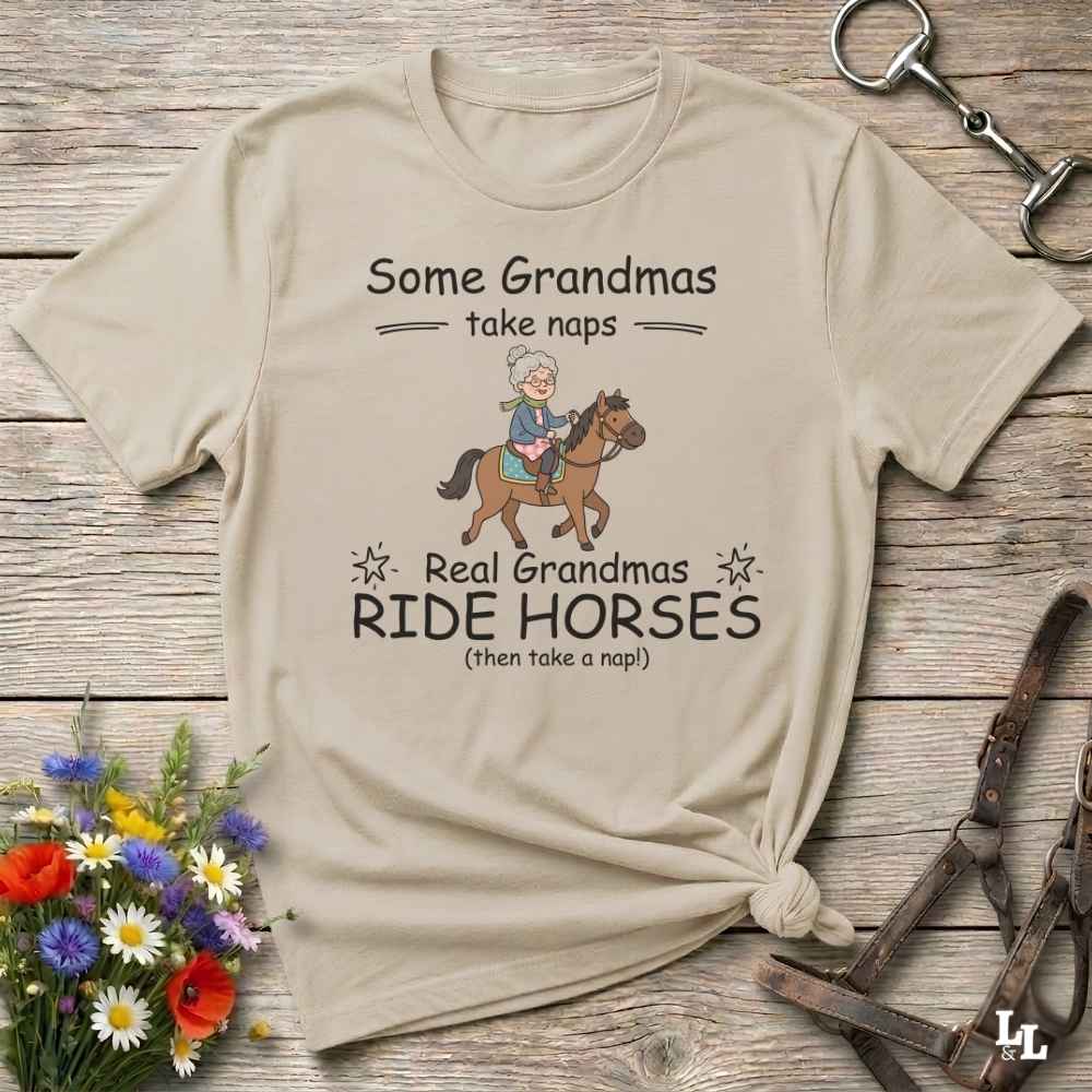 A beige graphic t-shirt laid flat on a rustic wooden surface, styled with equestrian accessories and a small bouquet of flowers. The shirt features a cartoon illustration of a grandma riding a horse, with the text 'Some Grandmas take naps' and 'Real Grandmas RIDE HORSES (then take a nap!)'.