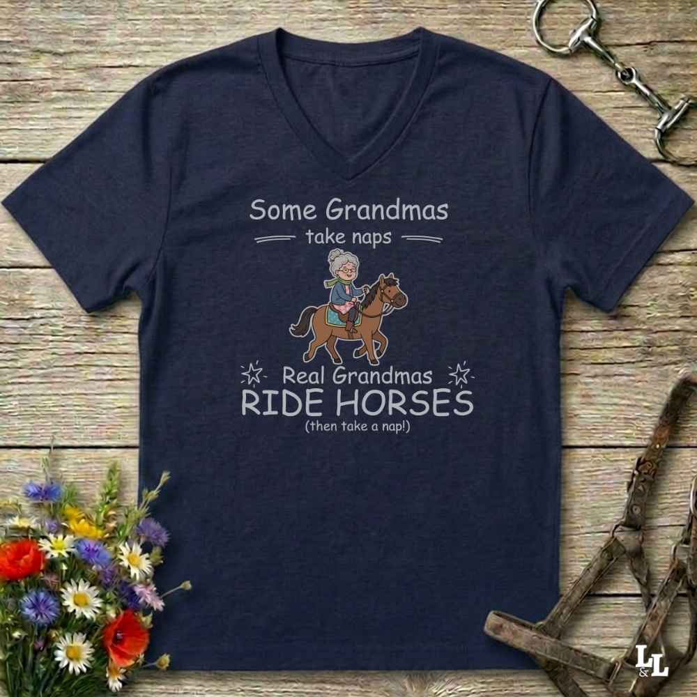 A navy blue V-neck t-shirt with a graphic print of a grandma riding a horse and the text 'Some Grandmas take naps Real Grandmas RIDE HORSES (than take a nap)'. The shirt is displayed in a flat-lay arrangement on a rustic wooden background, surrounded by decorative props including a flower bouquet, a horse bit, and a halter.