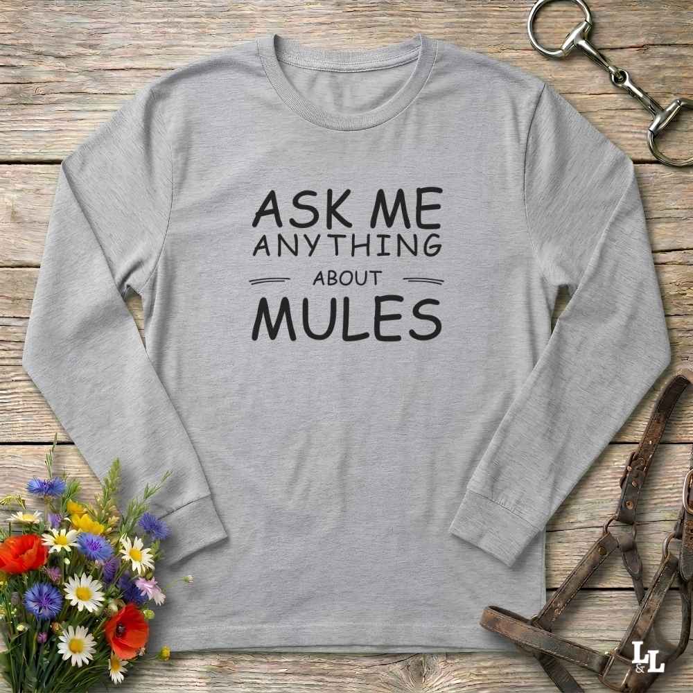 A light gray long-sleeve t-shirt with the text 'ASK ME ANYTHING ABOUT MULES' printed in black on the front, displayed in a flat-lay arrangement on a rustic wooden background with equestrian-themed props.