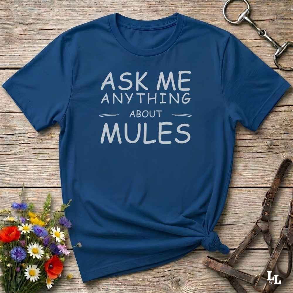 A navy blue graphic t-shirt with the text 'ASK ME ANYTHING ABOUT MULES' in white, styled in a flat-lay on a rustic wooden background with flowers and horse tack.