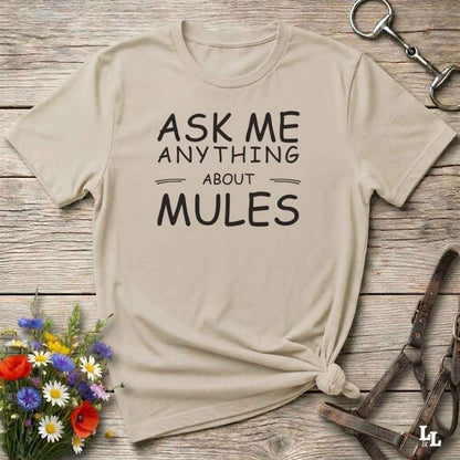 A beige graphic t-shirt with the text 'ASK ME ANYTHING ABOUT MULES' printed in black, styled in a flat-lay on a rustic wooden background with equestrian accessories and flowers.