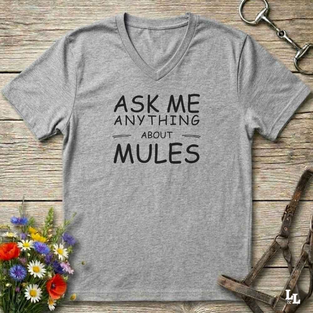A heather gray V-neck t-shirt with black text reading 'ASK ME ANYTHING ABOUT MULES' laid flat on a rustic wooden surface, styled with flowers, a horse bit, and a horse halter.