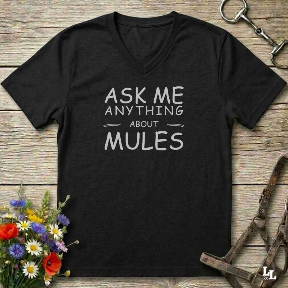 A black V-neck t-shirt with the text 'ASK ME ANYTHING ABOUT MULES' printed in white, laid flat on a rustic wooden background with equestrian-themed props.