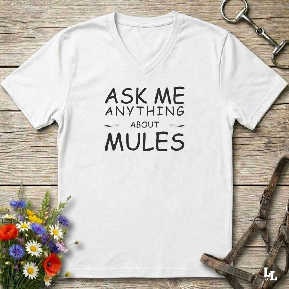 A white V-neck t-shirt with black text reading 'ASK ME ANYTHING ABOUT MULES' laid flat on a rustic wooden surface, styled with flowers, a horse bit, and a leather strap.