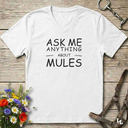A white V-neck t-shirt with black text reading 'ASK ME ANYTHING ABOUT MULES' laid flat on a rustic wooden surface, styled with flowers, a horse bit, and a leather strap.