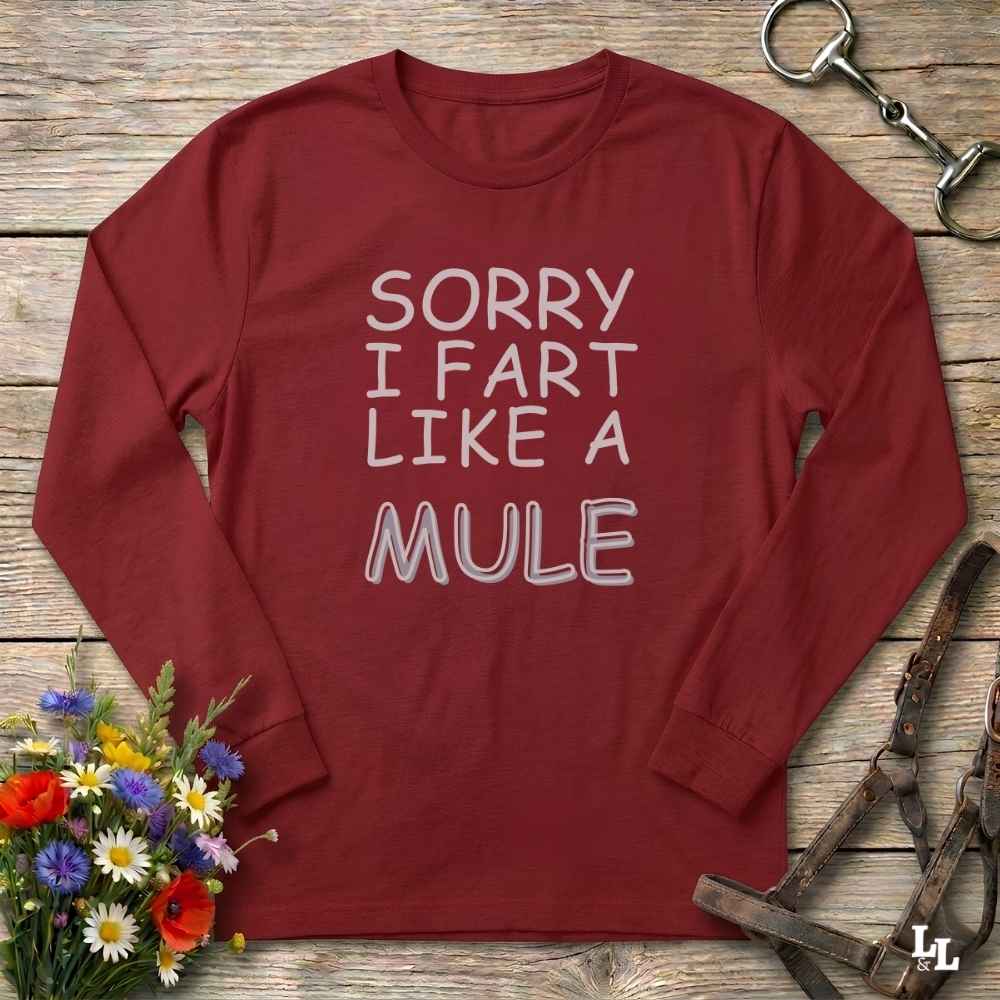 A long-sleeve crewneck t-shirt in maroon red with the text 'SORRY I FART LIKE A MULE' printed in white across the chest. The shirt is displayed flat on a rustic wooden background, styled with equestrian tack (a bit and bridle) and a small bouquet of colorful wildflowers.
