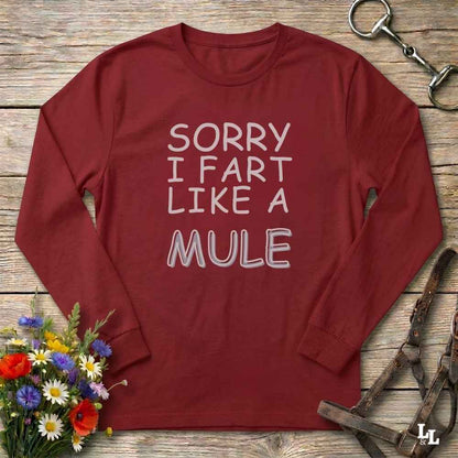 A long-sleeve crewneck t-shirt in maroon red with the text 'SORRY I FART LIKE A MULE' printed in white across the chest. The shirt is displayed flat on a rustic wooden background, styled with equestrian tack (a bit and bridle) and a small bouquet of colorful wildflowers.