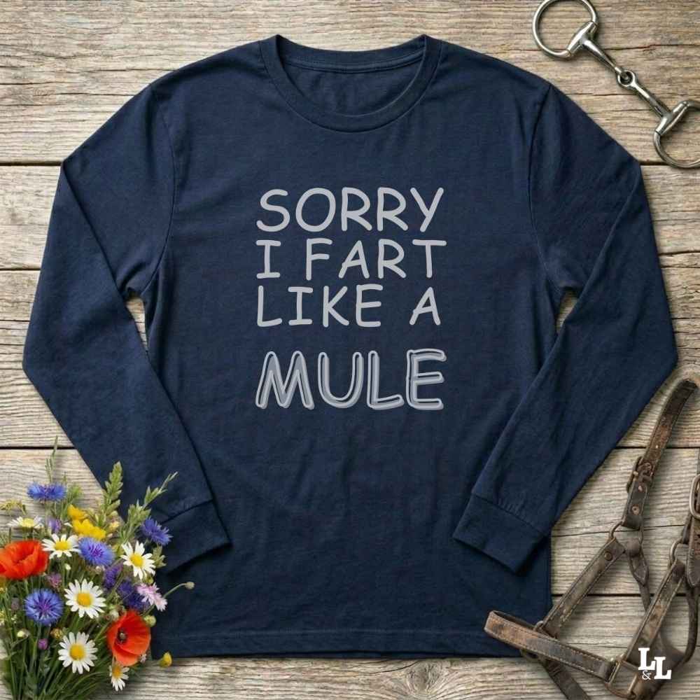 A navy blue long-sleeve t-shirt with the text 'SORRY I FART LIKE A MULE' printed in white on the front, styled in a flat-lay on a rustic wooden background with equestrian accessories and flowers.