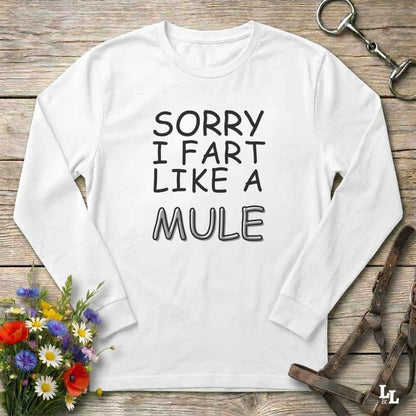 A white long-sleeve t-shirt with the text 'SORRY I FART LIKE A MULE' printed in black on the front, laid flat on a rustic wooden background with decorative horse tack and flowers.
