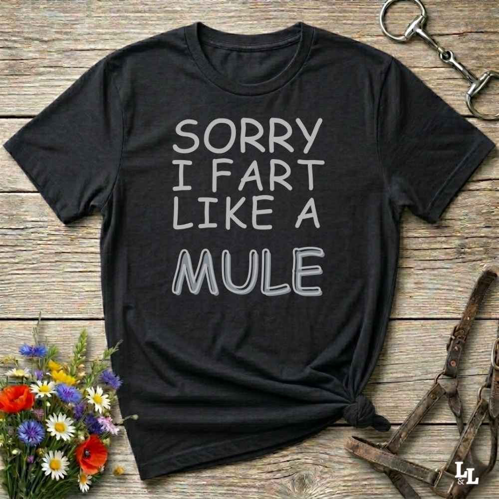 A black graphic t-shirt with the text 'SORRY I FART LIKE A MULE' printed in white, styled in a flat-lay on a rustic wooden background with equestrian accessories and flowers.