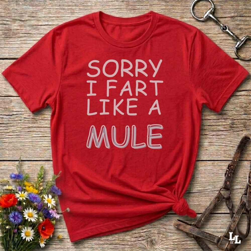 A red short-sleeve graphic T-shirt laid flat on a rustic wooden surface, with white text reading “SORRY I FART LIKE A MULE.” Decorative flowers and equestrian tack are placed around it as styling props.