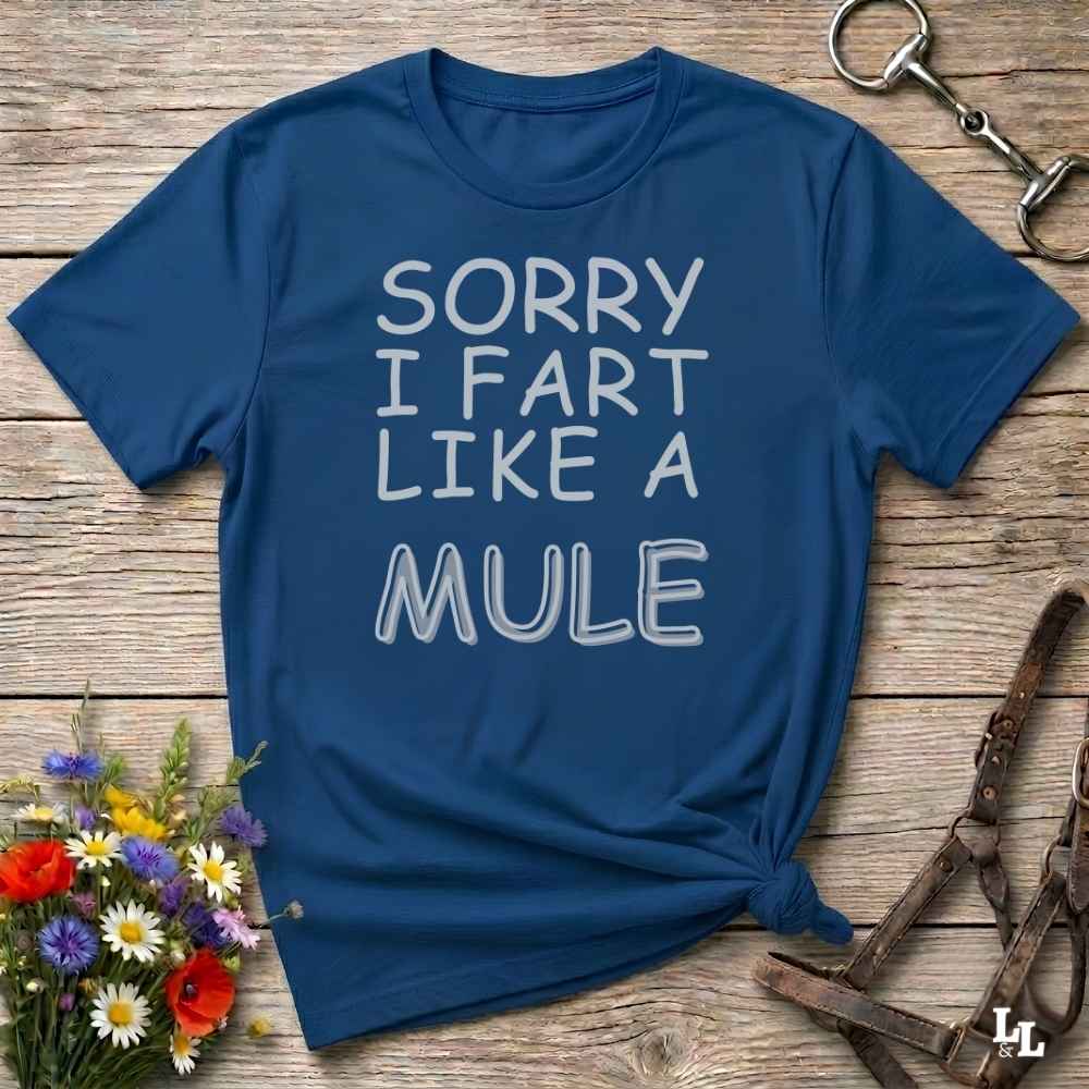 A navy blue short-sleeve t-shirt with the text 'SORRY I FART LIKE A MULE' printed in white across the front, styled in a flat-lay on a rustic wooden background with equestrian accessories and flowers.