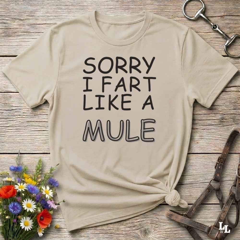 A beige graphic t-shirt with the text 'SORRY I FART LIKE A MULE' printed in black, styled in a flat-lay on a rustic wooden background with decorative flowers and leather tack.