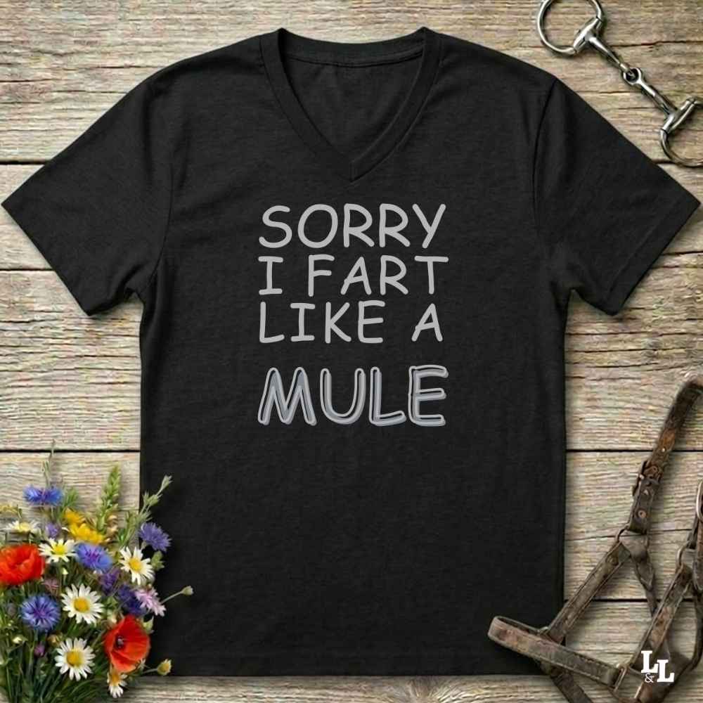 A black V-neck t-shirt with the text 'SORRY I FART LIKE A MULE' printed in white and gray outline lettering, displayed on a rustic wooden background with equestrian accessories and flowers.