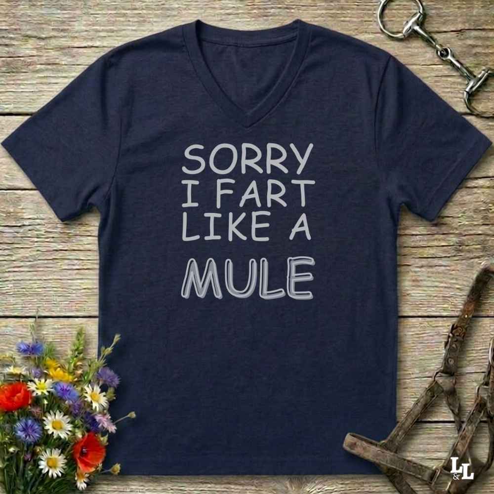 A navy blue V-neck t-shirt with the text 'SORRY I FART LIKE A MULE' printed in white outline lettering, laid flat on a rustic wooden background with equestrian-themed props.