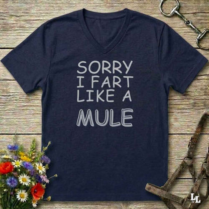 A navy blue V-neck t-shirt with the text 'SORRY I FART LIKE A MULE' printed in white outline lettering, laid flat on a rustic wooden background with equestrian-themed props.