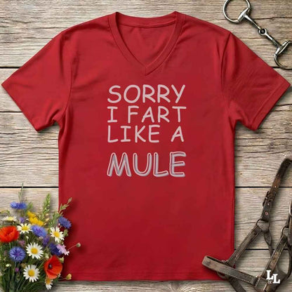 A red V-neck t-shirt with white text reading 'SORRY I FART LIKE A MULE' laid flat on a rustic wooden surface, styled with equestrian tack and a small bouquet of flowers.