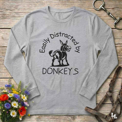 A light heather gray long-sleeve t-shirt with a black graphic print of a donkey and the text 'Easily Distracted by DONKEYS' laid flat on a rustic wooden surface with decorative equestrian and floral props.