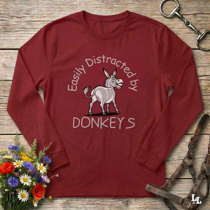 A long-sleeve crewneck t-shirt in maroon red, laid flat on a rustic wooden background. The shirt features a graphic of a donkey with the text 'Easily Distracted by DONKEYS' in white.