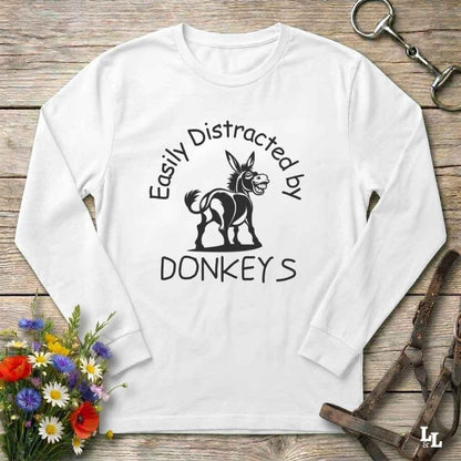 A white long-sleeve t-shirt with a black graphic print of a donkey and the text 'Easily Distracted by DONKEYS', displayed flat on a rustic wooden background with equestrian accessories and flowers.