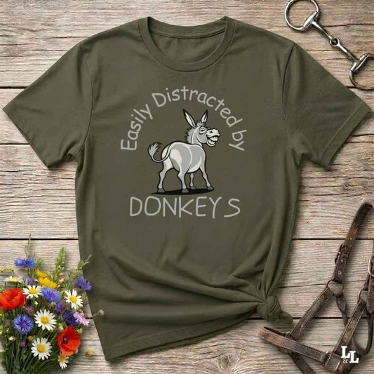 An olive green graphic t-shirt laid flat on a rustic wooden surface, styled with equestrian accessories and a small bouquet of flowers. The shirt features a cartoon donkey and the text 'Easily Distracted by DONKEYS'.