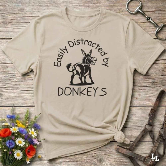 A beige graphic t-shirt with a black donkey illustration and the text 'Easily Distracted by DONKEYS', styled in a flat-lay on a rustic wooden background with flowers and horse tack.