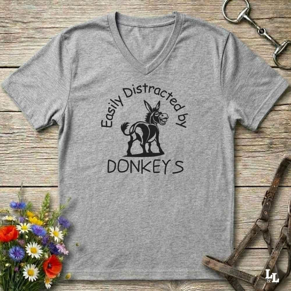 A heather gray V-neck t-shirt with a black graphic of a donkey and the text 'Easily Distracted by DONKEYS' laid flat on a rustic wooden background with decorative flowers and horse tack.