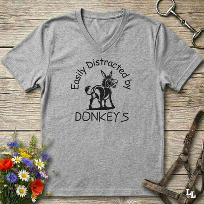 A heather gray V-neck t-shirt with a black graphic of a donkey and the text 'Easily Distracted by DONKEYS' laid flat on a rustic wooden background with decorative flowers and horse tack.