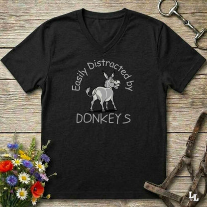 A black V-neck t-shirt with a white graphic of a donkey and the text 'Easily Distracted by DONKEYS' printed on the front, displayed on a rustic wooden background with equestrian accessories and flowers.