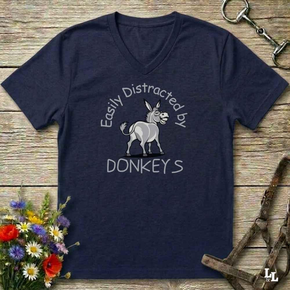 A navy blue V-neck t-shirt with a graphic of a donkey and the text 'Easily Distracted by DONKEYS' printed on the front, displayed on a rustic wooden background with equestrian accessories and flowers.