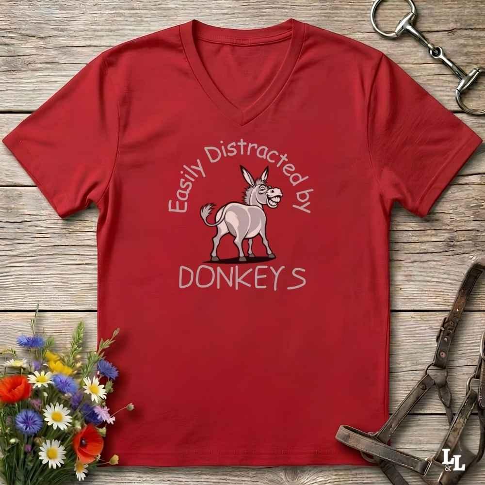 A red V-neck t-shirt with a graphic of a donkey and the text 'Easily Distracted by DONKEYS' printed on the front, displayed on a rustic wooden background with equestrian accessories and flowers.