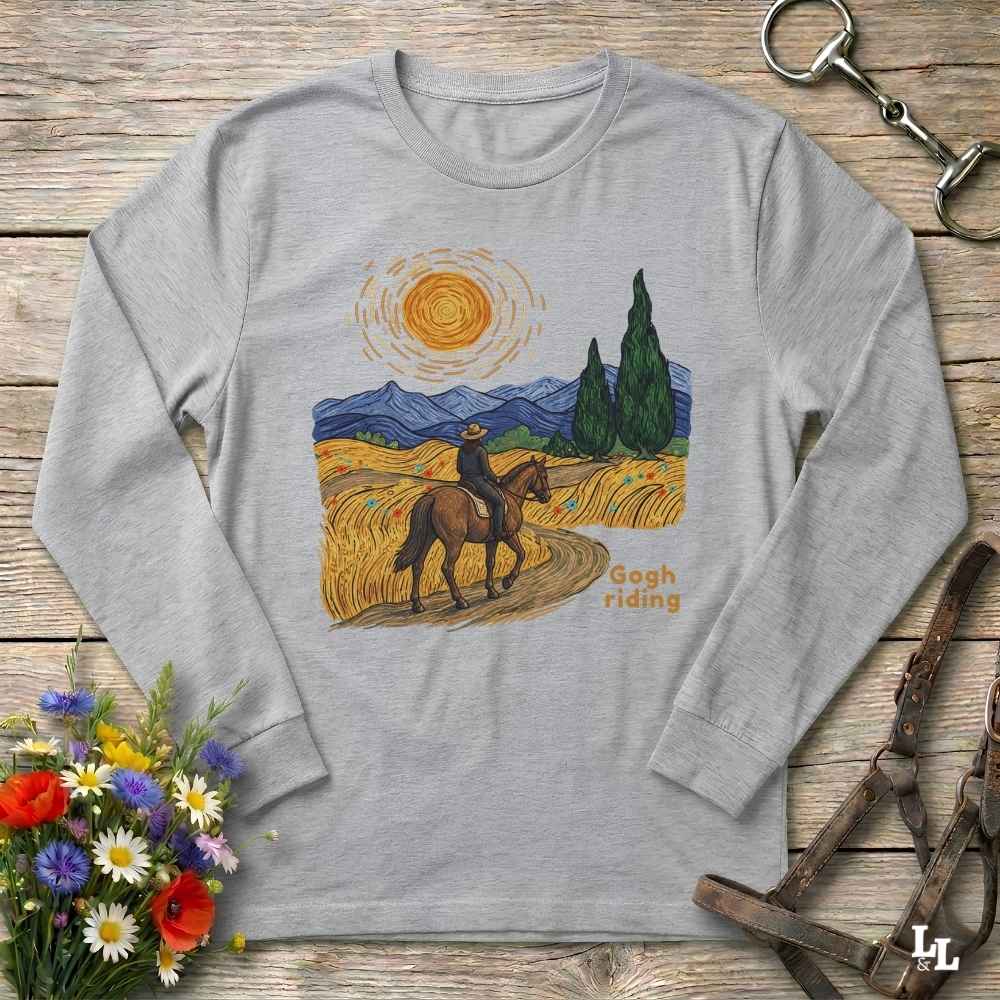 A light gray long-sleeve t-shirt with a colorful graphic print of a cowboy riding a horse through a field under a bright sun, with the text 'Sooth riding'. The shirt is displayed flat on a rustic wooden background with equestrian accessories and flowers.