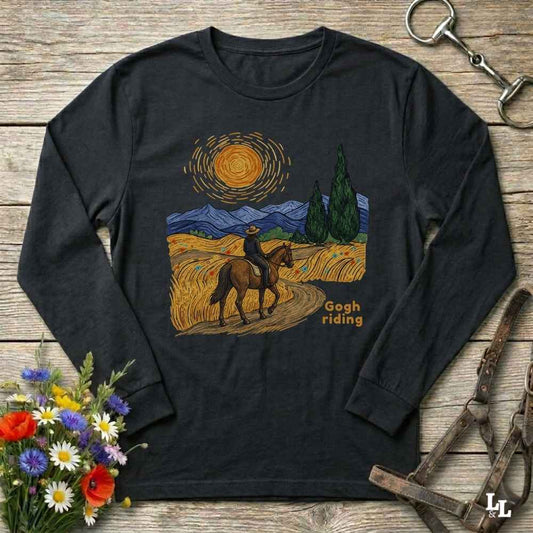 A black long-sleeve t-shirt with a colorful graphic print of a cowboy riding a horse through a stylized landscape in a Van Gogh-inspired style, with the text 'Gogh riding' printed below. The shirt is displayed flat on a rustic wooden background with decorative props including flowers and a leather bridle.