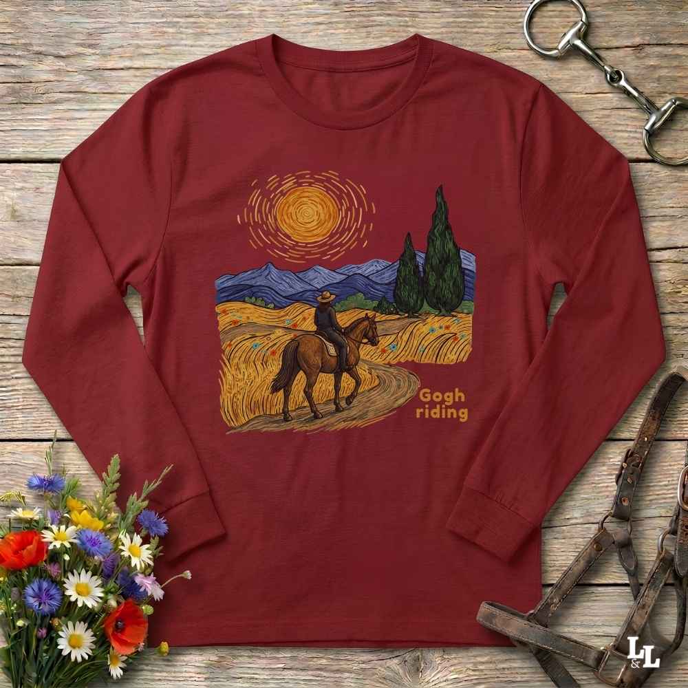 A long-sleeve crewneck t-shirt in maroon red, laid flat on a rustic wooden surface. The front of the shirt features a colorful graphic print depicting a cowboy riding a horse through a golden field under a swirling sun, with mountains and cypress trees in the background, and the text 'Gogh riding'.