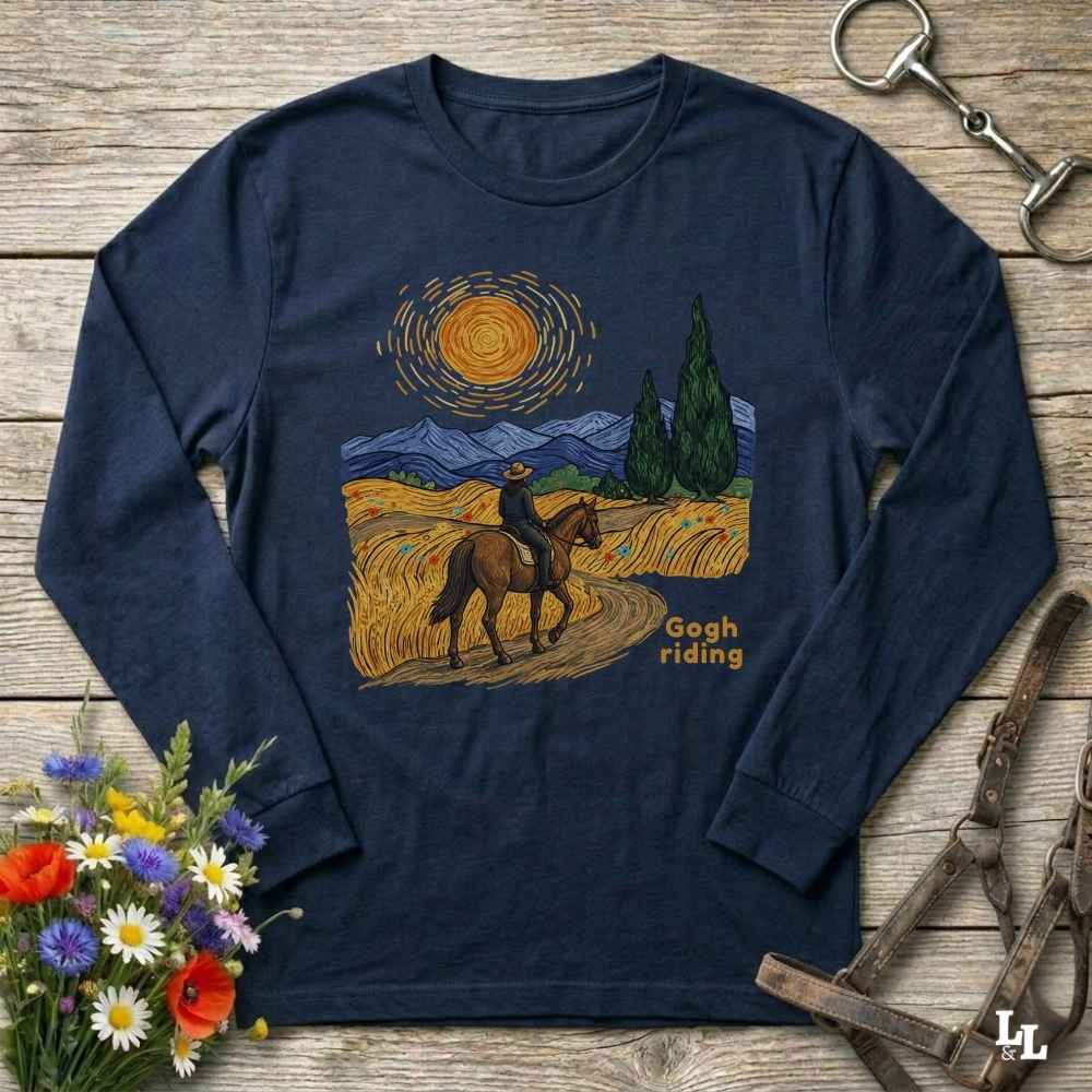 A navy blue long-sleeve t-shirt with a graphic print of a cowboy riding a horse in a stylized Van Gogh landscape, laid flat on a rustic wooden background with decorative flowers and horse tack.