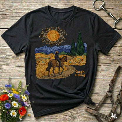 A black graphic t-shirt laid flat on a rustic wooden surface, styled with flowers, a horse bit, and a leather bridle. The t-shirt features a Van Gogh-inspired artwork of a rider on a horse through a golden field under a swirling yellow sun, with the text 'Gogh riding'.