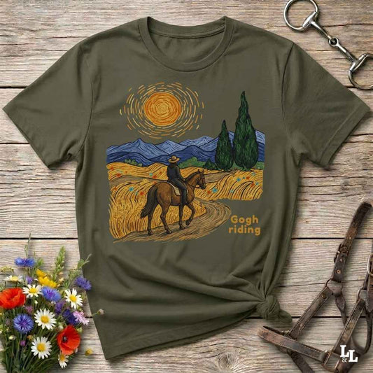 An olive green graphic t-shirt laid flat on a rustic wooden surface, styled with a horse bit, leather reins, and a small bouquet of wildflowers. The t-shirt features a Van Gogh-inspired print of a cowboy riding a horse through a golden wheat field under a swirling yellow sun, with blue mountains and green cypress trees in the background.