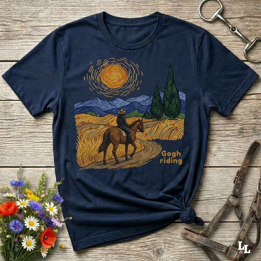 A navy blue graphic t-shirt laid flat on a rustic wooden surface, styled with flowers, a horse bit, and a leather bridle. The t-shirt features a graphic print of a rider on a horse in a field under a swirling yellow sun, styled like Van Gogh's Starry Night, with the text 'Gogh riding'.