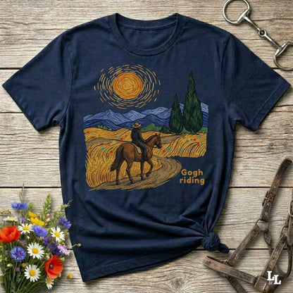 A navy blue graphic t-shirt laid flat on a rustic wooden surface, styled with flowers, a horse bit, and a leather bridle. The t-shirt features a graphic print of a rider on a horse in a field under a swirling yellow sun, styled like Van Gogh's Starry Night, with the text 'Gogh riding'.