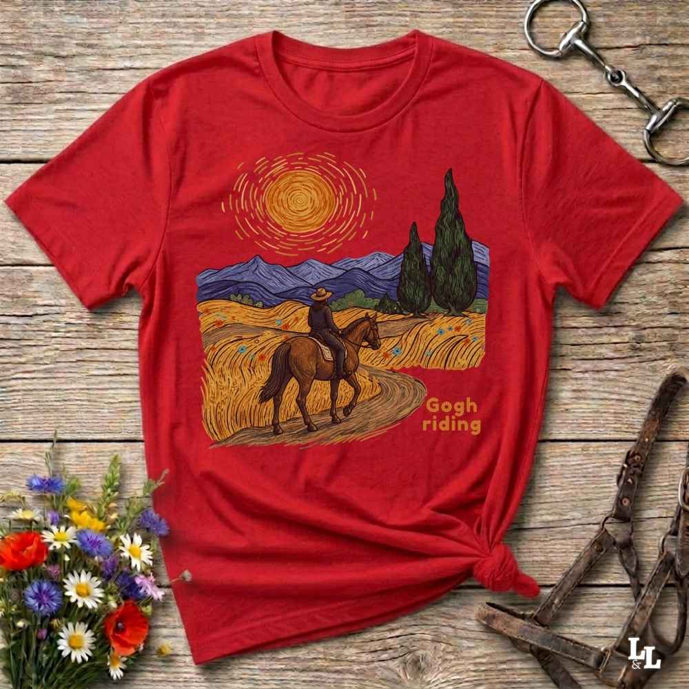 A red short-sleeve graphic T-shirt laid flat on a rustic wooden surface, styled with flowers, a horse bit, and a leather bridle. The shirt features a Van Gogh-inspired landscape print with a rider on horseback, blue mountains, cypress trees, and the words 'Gogh riding'.