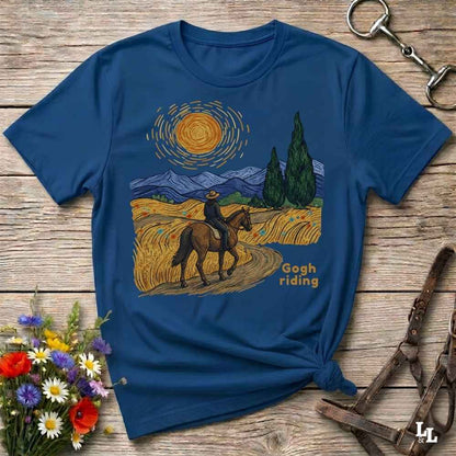 A navy blue graphic t-shirt laid flat on a rustic wooden surface, styled with equestrian and floral props. The shirt features a Van Gogh-inspired print of a rider on a horse through a golden field under a swirling yellow sun.